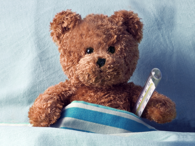 Teddy bear sitting in bed with a thermometer under its arm